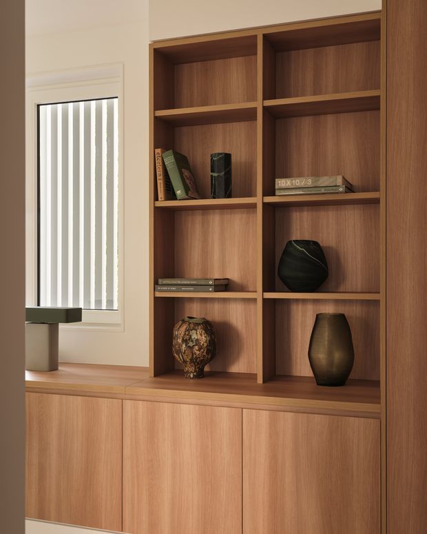 Close-up interior view of a built-in timber bookcase and lower storage cabinetry beside a window with vertical blinds. The warm wood joinery contains books and sculptural decorative objects, creating a refined, quiet display and storage feature.