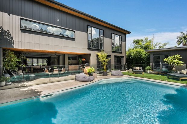 A contemporary two-storey dark-clad residence viewed from a landscaped backyard, featuring expansive glazing, a covered outdoor entertaining area, and a large turquoise swimming pool with surrounding lounge seating. A detached pavilion or pool house is visible to the right, with tropical and subtropical planting around the property.