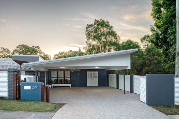 Contemporary single-storey residential facade with a broad skillion roof and covered carport/entry. The house uses white and dark grey rendered surfaces, horizontal slatted fencing, a brick letterbox, and a textured driveway, set among established trees.