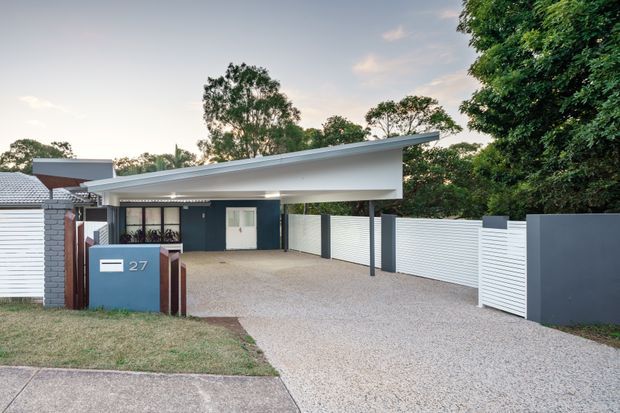Street-facing exterior of a contemporary single-storey residence with a broad angled skillion roof and large open carport. The facade combines dark painted walls, white horizontal privacy fencing, a textured light driveway, and a blue-grey entry pillar marked with the house number. Mature trees surround the property.