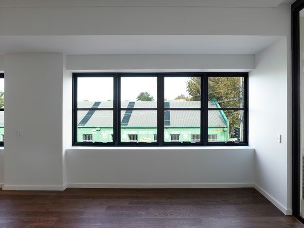 Bright, minimally furnished interior room with dark timber flooring, white walls and ceiling, and a wide black-framed horizontal window set into a recessed opening. The window overlooks neighbouring rooftops and greenery. A tall black-framed glazed opening is partially visible at the right edge.