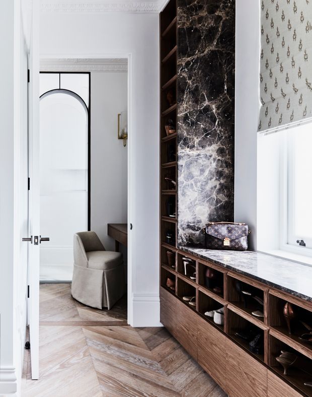 Refined transitional interior featuring a built-in dark timber storage and display wall with open shelving, drawers, and a dramatic dark veined marble feature slab and benchtop. The space has ornate white decorative mouldings, patterned Roman blinds, herringbone timber flooring, and a glimpse of an adjacent room with a curved black-framed doorway and upholstered chair.