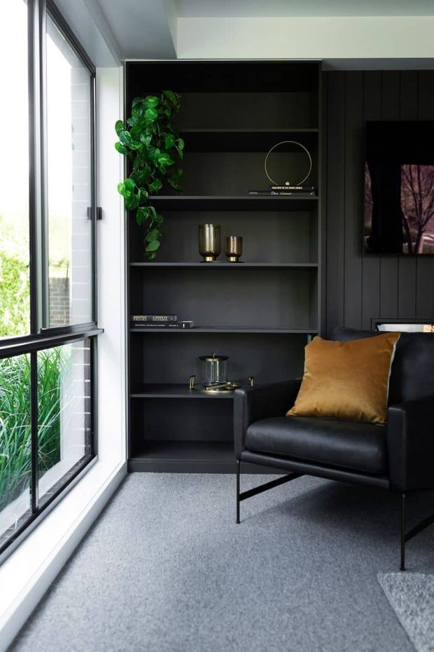 A refined living room corner with a full-height black built-in shelving unit, dark vertical wall panelling, a black leather armchair, and a mustard-gold cushion. Decorative books, brass-toned vessels, a circular ornament, and a trailing plant style the shelves. Large black-framed windows bring in abundant daylight and overlook greenery, while grey carpet provides a soft neutral base.