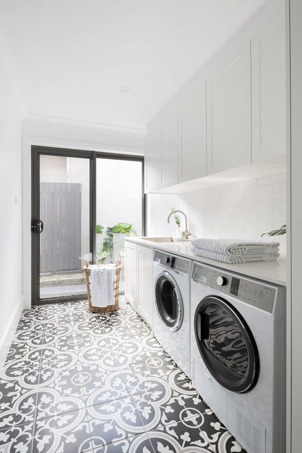Bright, compact contemporary laundry room with white built-in cabinetry, a white tiled splashback, integrated benchtop, front-loading washer and dryer, and ornate grey-and-white patterned floor tiles. A glazed exterior door provides natural light, with folded towels and a small basket adding practical detail.