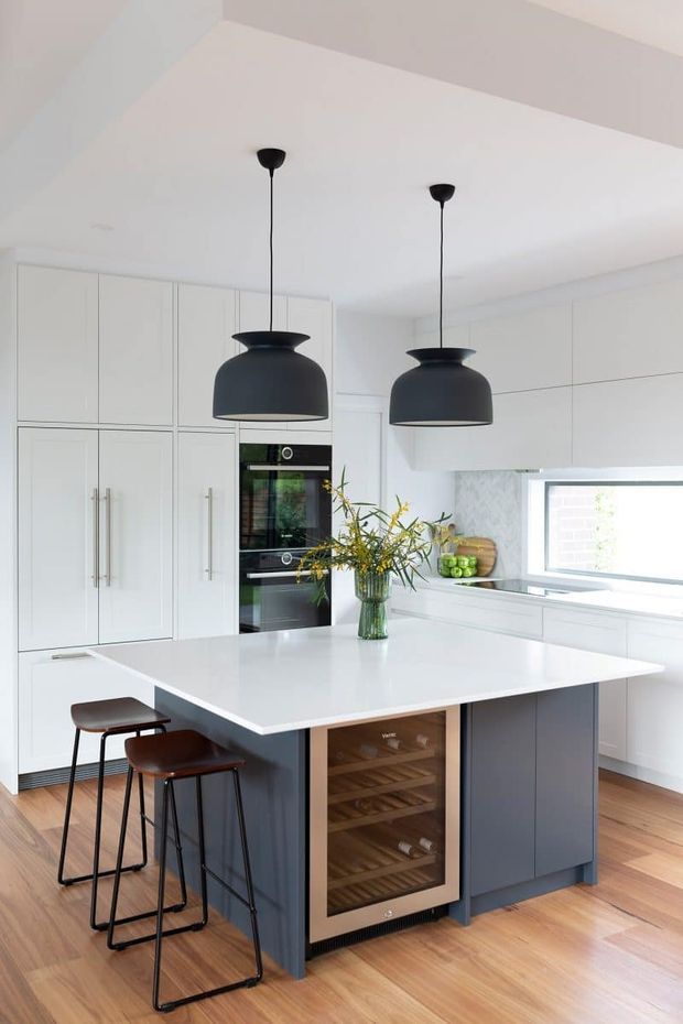Bright contemporary kitchen featuring a large white island with dark blue-grey cabinetry, integrated wine storage, white full-height cabinetry, built-in ovens, pendant lights, timber flooring, and a compact dining overhang with stools. The space has a clean, refined monochrome palette with warm timber accents.
