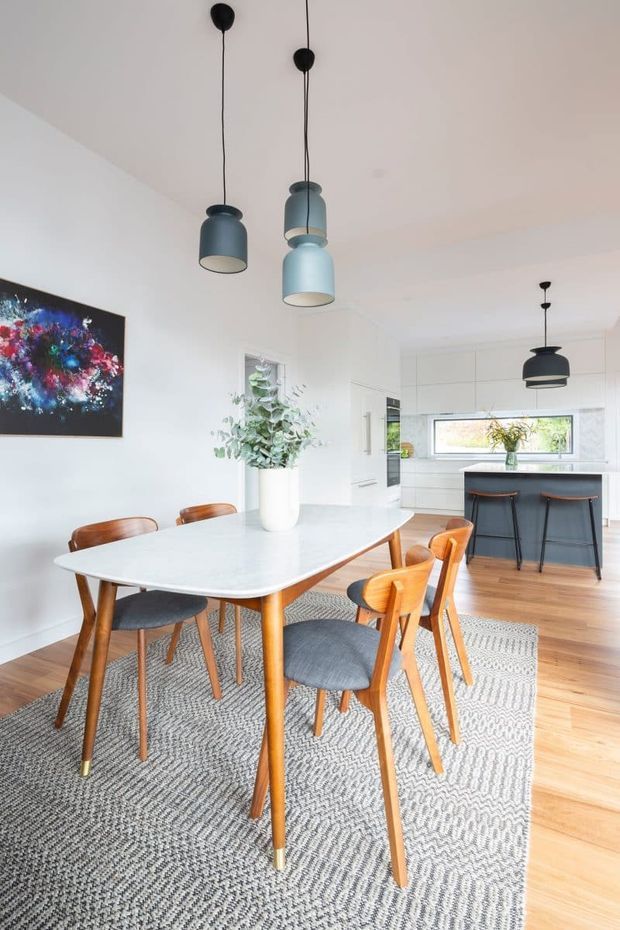 Bright open-plan dining area adjoining a contemporary kitchen. A white rounded rectangular dining table is surrounded by mid-century timber chairs with grey upholstered seats, set over a large patterned grey rug. Three blue-grey pendant lights hang above the table, while a black pendant accents the white kitchen and dark island benchtop in the background. Timber flooring, white cabinetry, a feature artwork, and indoor greenery complete the space.