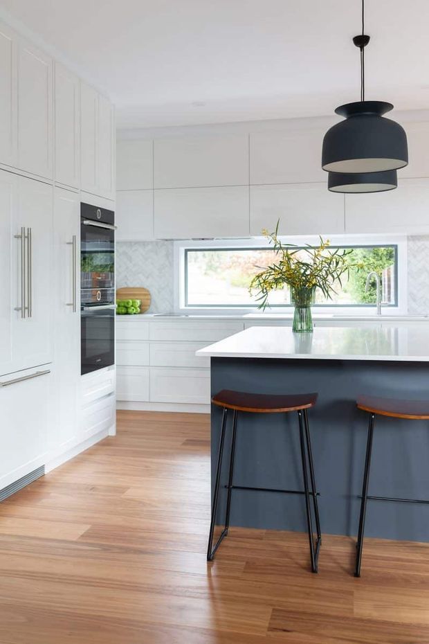 Bright contemporary kitchen with extensive white flat-panel cabinetry, integrated appliances, a large dark island with a white benchtop, timber bar stools, warm timber flooring, and a pair of oversized black pendant lights. A long horizontal window and pale patterned splashback add light and texture.