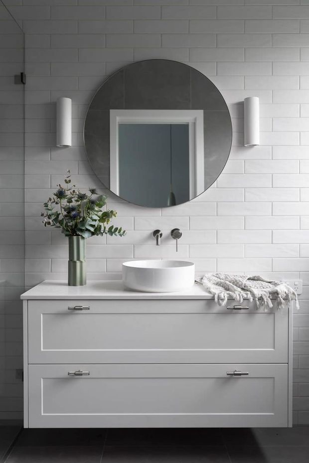 A refined contemporary bathroom vanity vignette featuring a white wall-mounted-style cabinet with shaker-profile drawers, a white vessel basin, wall-mounted chrome tapware, and a large round mirror. The backsplash is clad in white rectangular tiles with light grout, complemented by vertical white cylindrical wall lights and a muted floral arrangement. Dark floor tiles provide contrast.