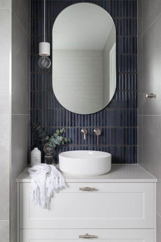 A compact, refined bathroom vanity featuring a white vessel basin on a pale grey stone benchtop, white paneled cabinetry, grey wall tiles, and a striking deep blue vertically ribbed tile splashback. A large rounded-arch mirror, wall sconce, brushed metal tapware, and understated accessories complete the contemporary scheme.