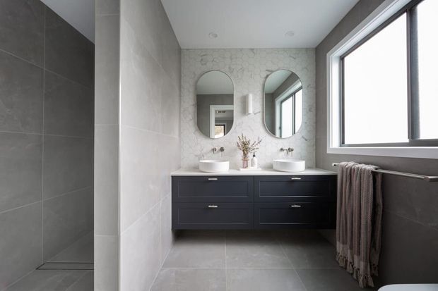 Contemporary double-vanity bathroom with dark floating cabinetry, twin white vessel basins, arched mirrors, pale veined wall tiles, large-format grey floor and wall tiles, and a wide black-framed window. The space is bright, clean and refined with a restrained neutral palette.