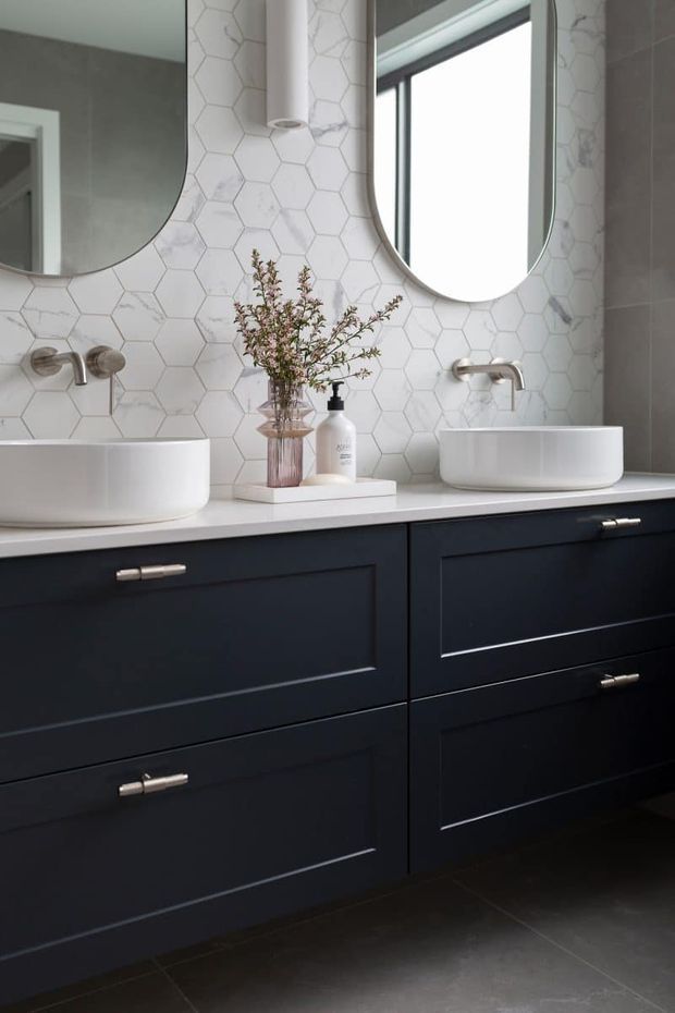 A refined contemporary bathroom vanity with two white vessel basins set on a pale benchtop, dark navy-blue shaker-style cabinetry, brushed metal wall-mounted taps, and rounded rectangular mirrors. The wall is finished with white marble-look hexagonal tiles, while large dark floor tiles and a narrow window complete the space. A small vase and soap dispenser provide restrained styling.