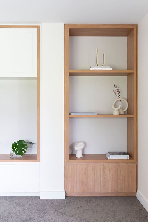 A bright, minimalist built-in shelving wall featuring warm timber open shelves and lower cupboards, white painted cabinetry and trim, and carefully styled decorative objects and books. A grey carpeted floor and a small potted tropical leaf add soft contrast.