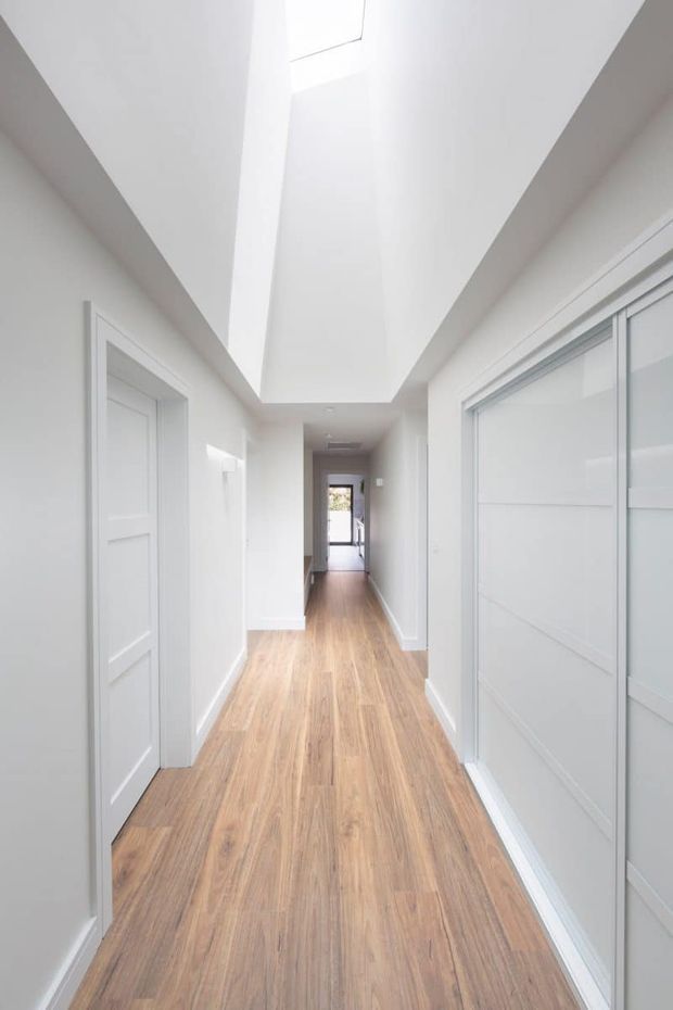 A bright, narrow residential hallway with white walls, white paneled doors and sliding built-in doors, warm timber-look flooring, and a dramatic elongated ceiling void or skylight that brings in natural light. The corridor leads toward a distant doorway.