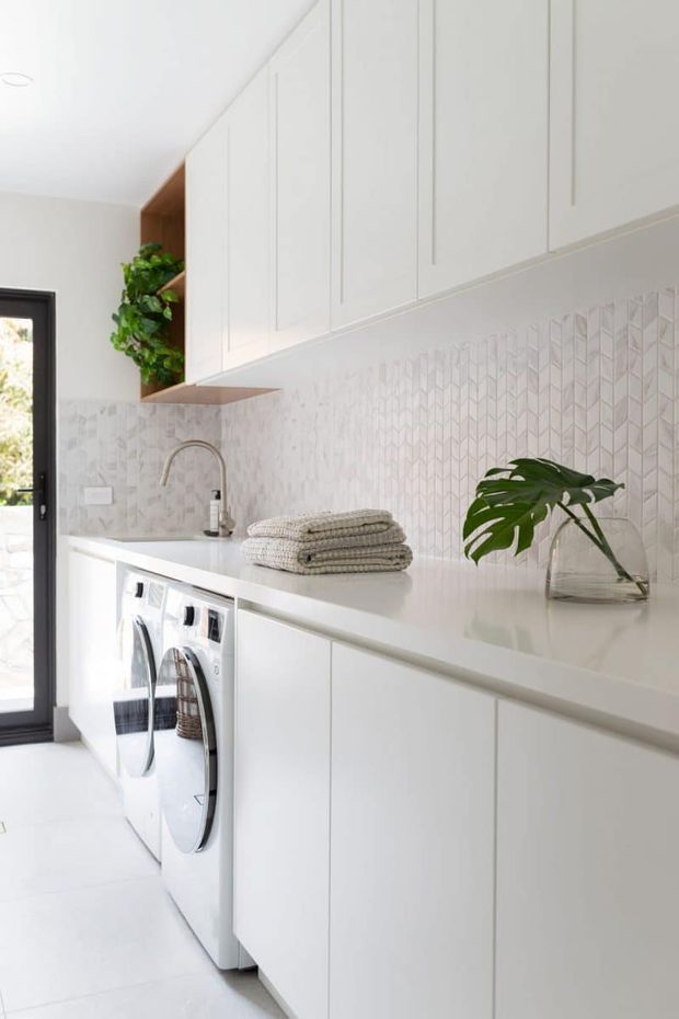 Bright, compact contemporary laundry room with white built-in cabinetry, a long white benchtop, front-loading washer and dryer, integrated sink with curved chrome tap, and a pale geometric tiled splashback. A dark-framed glazed door, folded towels, and indoor plants add contrast and warmth.