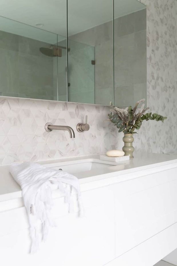 A bright, refined contemporary bathroom featuring a white floating vanity with integrated basin, wall-mounted brushed metal tapware, a large mirrored cabinet, and a glass shower enclosure. The wall behind the vanity is finished with pale geometric textured tiles, while a soft white palette and muted decorative vase create a calm, minimalist atmosphere.