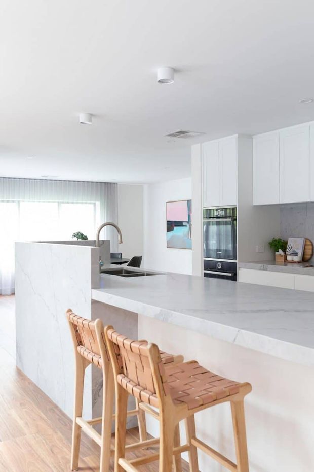 Bright, contemporary white kitchen with a large waterfall-edge island and integrated breakfast bar. The island has a pale veined stone surface, white cabinetry, a built-in oven, sink and curved brushed-metal tap. Two woven timber bar stools sit at the island. Floor-to-ceiling sheer curtains and a large window provide abundant natural light, with a colourful artwork on the rear wall.