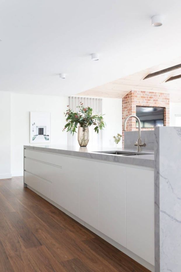 Bright contemporary kitchen featuring a long white island with integrated cabinetry, a grey stone benchtop, stainless steel undermount sink and curved mixer tap. The kitchen has warm timber flooring, a white ceiling with recessed spotlights, a pale timber-lined ceiling section, and an exposed red-brick fireplace wall with dark fireplace opening. A marble-look stone panel forms the island end panel, while sheer curtains and a floral arrangement add softness.