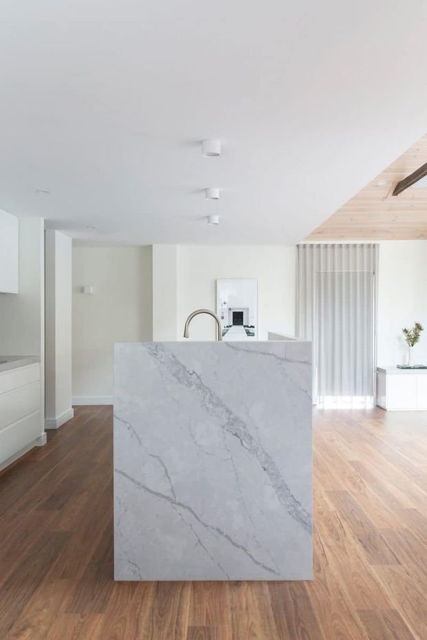 Bright open-plan contemporary kitchen and living area with a large white veined stone island, integrated cabinetry, dark timber flooring, white walls and ceiling, recessed lighting, and vertical sheer window coverings. A timber-lined vaulted ceiling section adds warmth at the right side of the room.