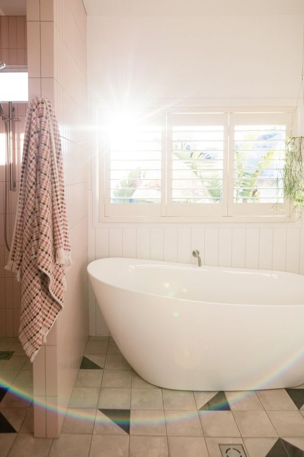 Bright, airy bathroom featuring a freestanding white bathtub beneath a wide white-framed louvred window. Pale vertical wall panelling, tiled flooring with small dark geometric accents, and a partially visible tiled shower area create a clean coastal feel. A patterned towel hangs beside the tub and a trailing plant is visible near the window.