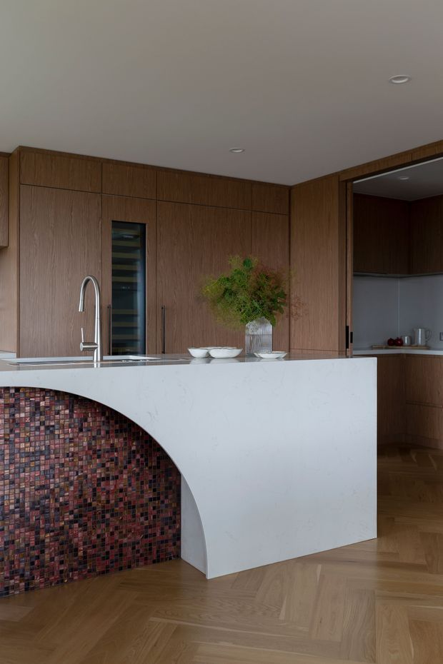 A refined contemporary kitchen with full-height warm timber cabinetry, a large white stone island, integrated sink and curved feature end clad in small multicoloured mosaic tiles. The space has a herringbone timber floor, recessed ceiling lights, dark timber accents, and a pantry or secondary kitchen area visible through an opening on the right.
