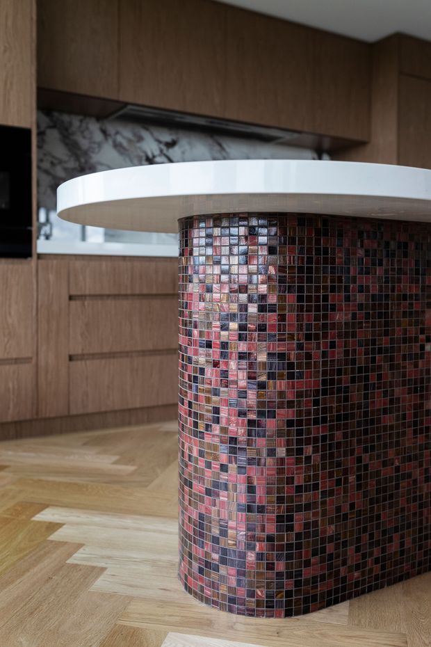 Close-up view of a contemporary kitchen featuring a curved island with a white rounded benchtop and a cylindrical base clad in small glossy mosaic tiles in burgundy, pink, brown, grey, and dark tones. The background includes warm timber cabinetry, a marble-look splashback, and light timber herringbone flooring.