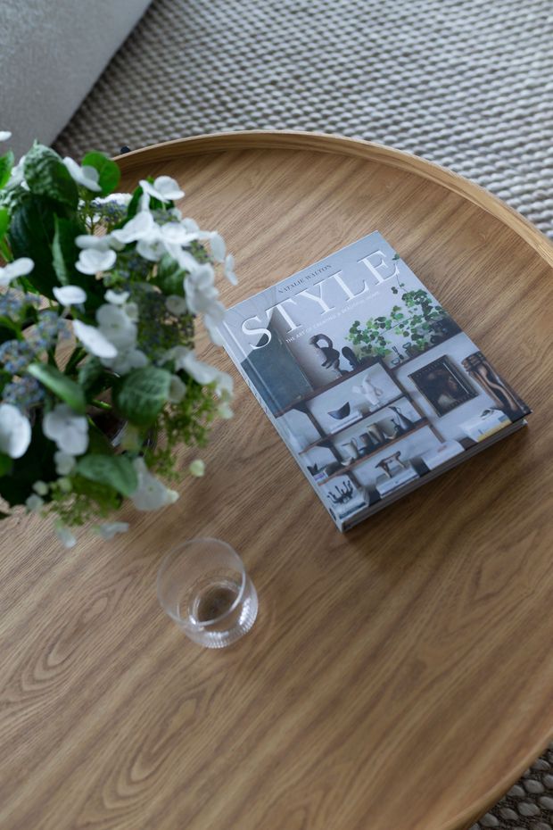 Close-up styled still life of a light timber oval coffee table with a raised rim, placed on a textured neutral rug. A design magazine, small glass, and leafy flowering plant sit on the tabletop.