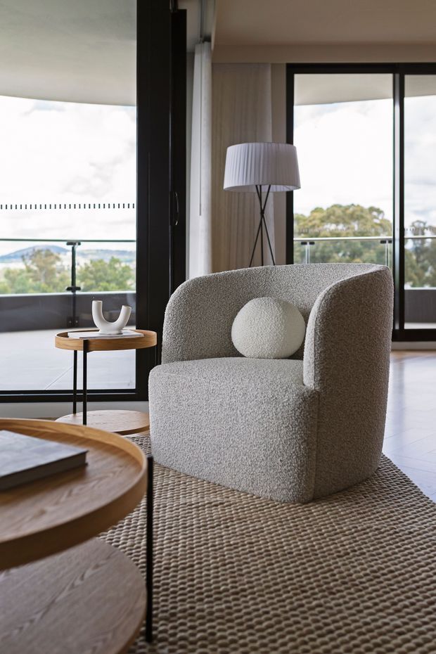 A refined contemporary living room with a textured boucle armchair, round light-coloured cushion, round timber coffee tables, woven rug, sculptural side table decor, and a floor lamp. Full-height black-framed glazing opens to a balcony with a glass balustrade and leafy views beyond. The room has a warm neutral palette and abundant natural light.