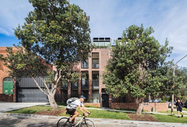 Street-facing view of a contemporary multi-storey brick residential building, partially screened by mature trees. The facade features dark-framed vertical windows, recessed balconies, rooftop elements, and a ground-level gated driveway beside neighbouring brick homes. A cyclist and pedestrian are visible on the street.