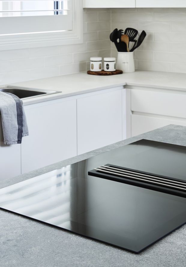 Bright, clean white kitchen corner with shaker-style cabinetry, pale benchtops, white subway-tile splashback, stainless-steel sink, and a large black glass induction cooktop set into the foreground island. A utensil holder and small canisters sit on the rear benchtop.