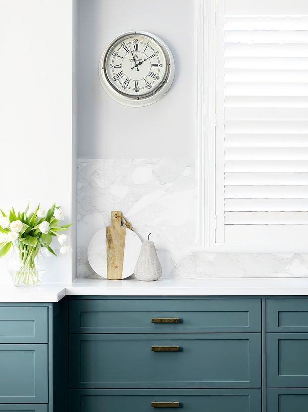 Bright, refined interior vignette showing a dark teal-blue built-in cabinet with shaker-style drawers and brass hardware, a white marble-look backsplash and benchtop, white plantation shutters, and a pale grey wall with a round wall clock. Decorative cutting boards, a ceramic vessel, and flowers add a soft traditional touch.