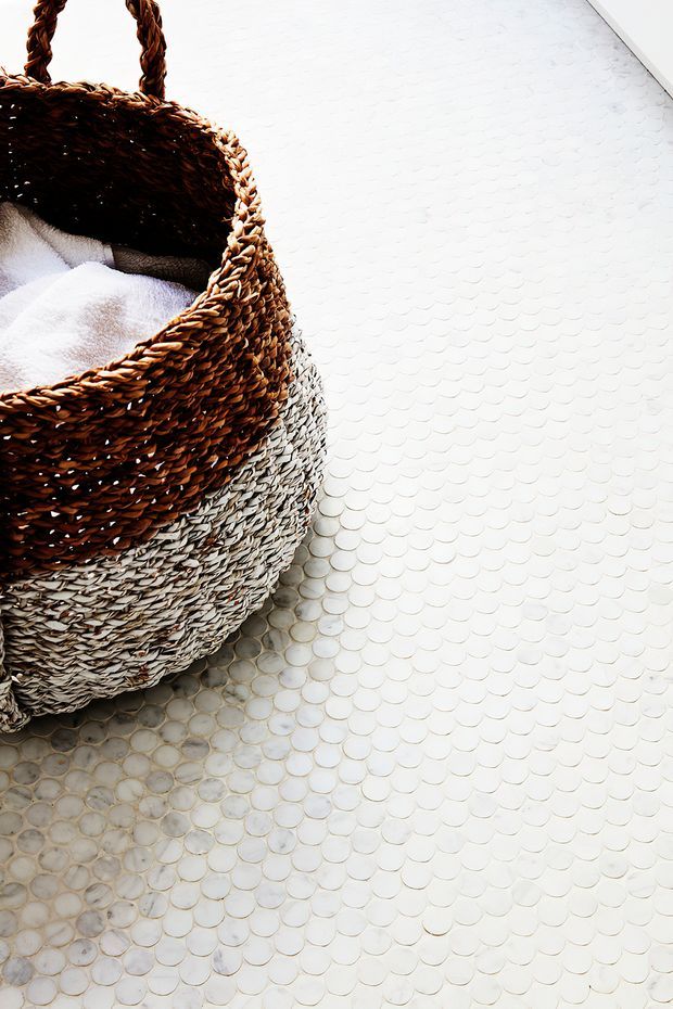 Close-up interior detail showing a woven wicker basket with a soft white towel, resting on a white circular mosaic tile floor. The image is bright and textural, with natural tan and grey basket fibres contrasting against the pale flooring.