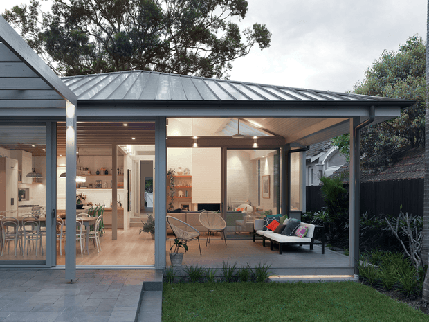 A contemporary single-storey home extension viewed from the garden, with a broad grey metal roof, dark-framed floor-to-ceiling glazing, and a covered outdoor living area. The glazed interior reveals an open-plan kitchen and dining space with light timber finishes, while the covered terrace contains lounge seating and connects directly to a small landscaped lawn and paved area.