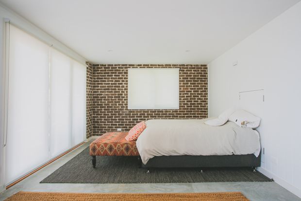 Bright contemporary bedroom with a low-profile upholstered bed, white bedding, patterned upholstered bench, large rug, white walls, broad roller blinds, and an exposed brick feature wall with a recessed window or opening.