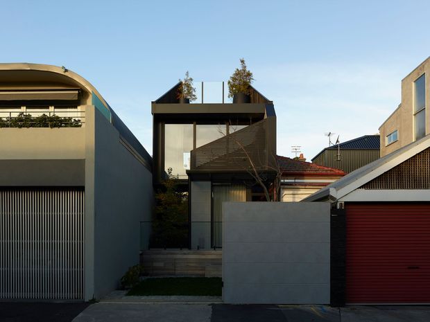 Street-facing view of a compact contemporary urban residence or townhouse composition. The facade features dark metal framing, large upper-level glazing, a projecting balcony with horizontal screening, a grey rendered or panelled boundary wall, and an integrated red roller garage door. Adjacent homes and narrow site spacing create a dense suburban streetscape.