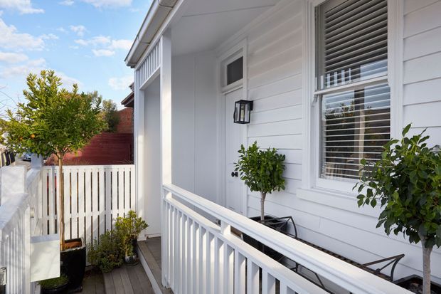 Bright exterior view of a white weatherboard-clad house featuring a covered porch or balcony, white timber balustrades, a white entry door, horizontal louvred blinds, and potted citrus trees. A red timber fence and neighbouring roof are visible in the background.
