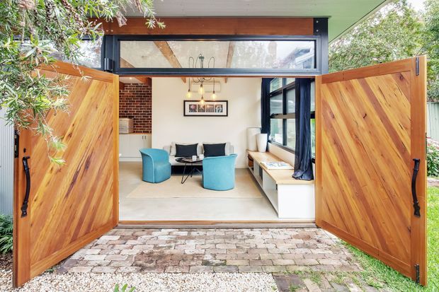 A bright, compact living room viewed from a landscaped courtyard through wide-open timber double doors. The space features white walls, pale flooring, a built-in window seat, black-framed glazing, a small sofa, turquoise lounge chairs, and a high horizontal clerestory window. Exposed brickwork and timber framing add warmth, while the exterior threshold is paved with reclaimed-looking brick and gravel.