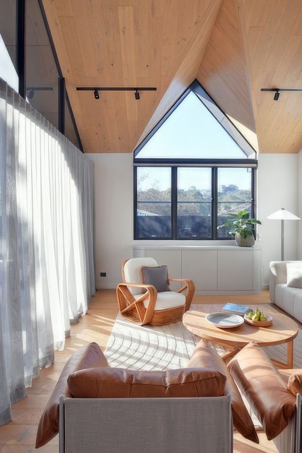 Bright, warm living room with a distinctive timber-lined vaulted ceiling and triangular clerestory window above a black-framed glazed window. Full-height sheer curtains line one side, while a leather sofa, curved timber lounge chair, round timber coffee table, floor lamp and built-in cabinetry create a comfortable contemporary interior. Sunlight casts strong linear shadows across the timber floor and rug.