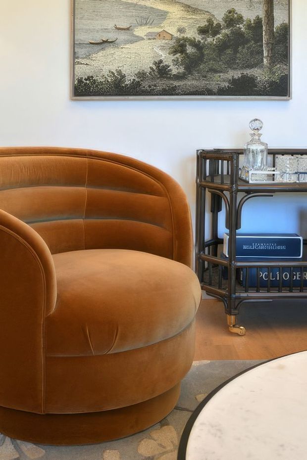 A warmly styled living room vignette featuring a large caramel-brown upholstered barrel chair with curved channel detailing, a dark wood or metal-framed bar cart with glassware, a framed coastal landscape artwork, timber flooring, and the edge of a round pale coffee table. The composition has a refined vintage character with warm, tactile materials and curved forms.