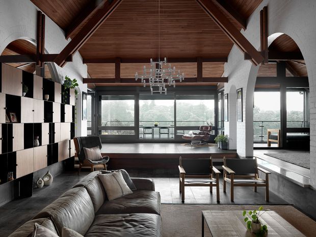 A spacious double-height living room with a dramatic exposed timber vaulted ceiling, dark structural beams, white textured masonry walls, extensive glazing, and a raised window-side seating area. The room contains a large sectional sofa, mid-century-style lounge chairs, modular dark built-in shelving, a sculptural chandelier, and a neutral textured rug. Lush greenery is visible beyond the glazing.