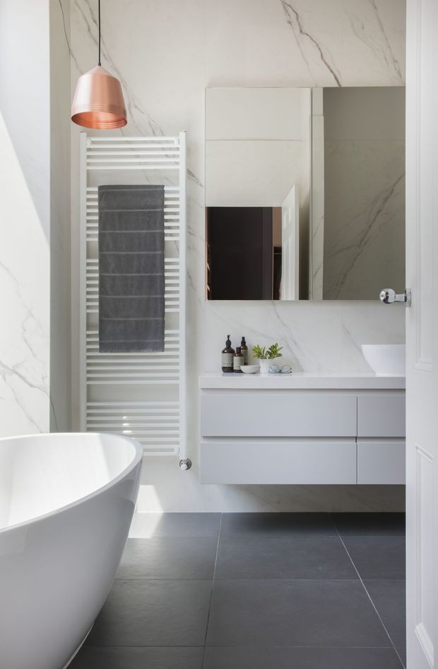 A bright, contemporary bathroom featuring large white marble-look wall tiles with grey veining, dark grey floor tiles, a white freestanding bathtub, wall-mounted pale grey vanity cabinetry, and a large mirrored cabinet. A white heated towel rail holds a dark grey towel, while a copper pendant light adds a warm accent.
