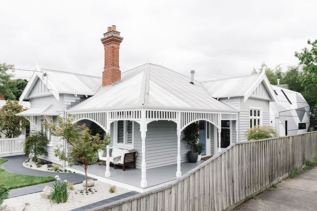 Exterior view of a single-storey white weatherboard house with multiple steeply pitched corrugated metal gables, decorative verandah arches and fretwork, louvred shutters, and a prominent red brick chimney. The home sits behind a timber boundary fence with landscaped paths and planting.