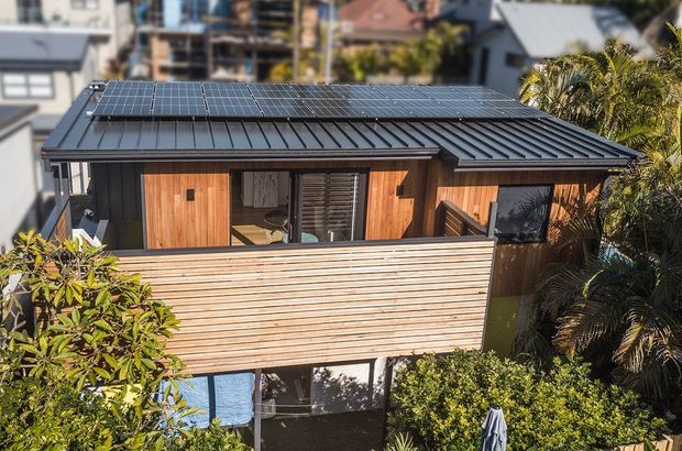 Elevated exterior view of a compact contemporary timber-clad house surrounded by dense greenery. The building has a dark standing-seam metal gable roof fitted with solar panels, warm vertical timber wall cladding, a horizontal timber privacy screen or balcony balustrade, and large glazed openings with external louvres. Parts of neighbouring urban housing are visible in the background.