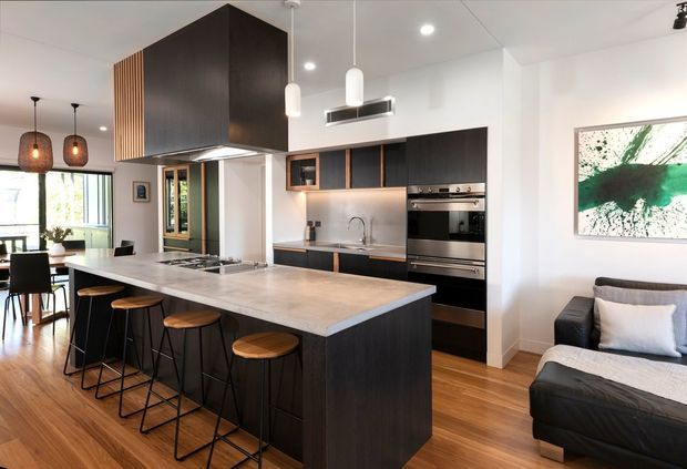 A bright, open-plan contemporary kitchen and living area. The kitchen features a large island with waterfall-style light stone benchtop, dark timber cabinetry, integrated stainless-steel appliances, a metallic splashback, pendant lights, and a prominent dark rangehood enclosure. A dining area is visible beyond the island, while a compact living area with a dark sofa and cushions occupies the right side. Timber flooring runs throughout.