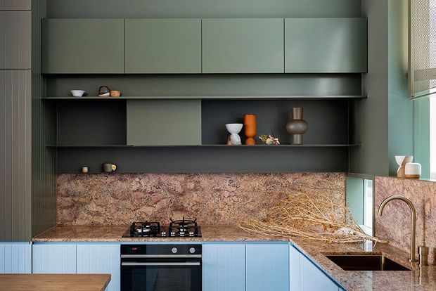 A contemporary kitchen with a compact L-shaped layout, muted blue vertically fluted lower cabinetry, dark charcoal open shelving and overhead cupboards, and a richly patterned stone backsplash and benchtop. The space includes a built-in oven, gas cooktop, undermount sink with a tall curved tap, and restrained decorative objects on the shelves. Natural light enters from the right through a window with a woven blind.