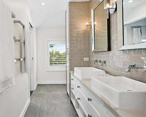 Bright contemporary bathroom with a long white double vanity, two square vessel basins, chrome wall-mounted taps, large mirrored cabinets, and a freestanding bathtub partially visible behind a glass shower screen. Glossy rectangular beige-grey wall tiles contrast with light grey floor tiles, while a shuttered window provides natural light.