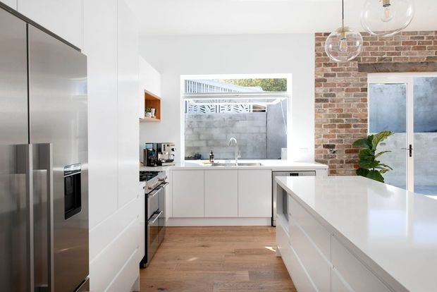 Bright contemporary kitchen with white flat-panel cabinetry, a large white island, stainless-steel appliances, timber flooring, and a red brick feature wall. A rear window provides natural light, while globe pendant lights hang above the island.