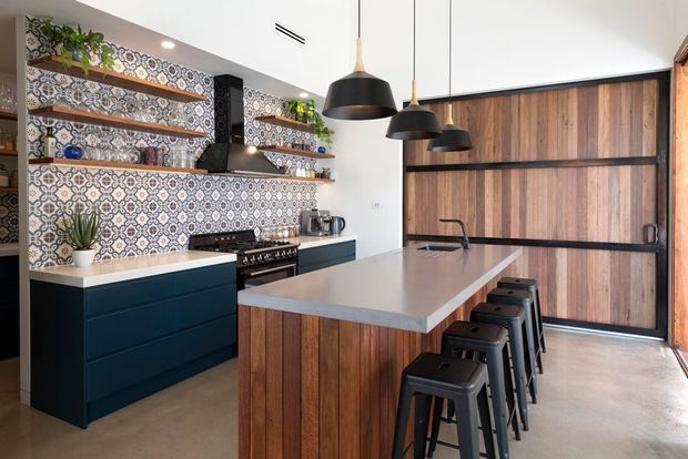 A contemporary kitchen with a large central island clad in vertical mid-toned timber, a pale stone benchtop, dark teal cabinetry, patterned geometric tile splashback, open timber shelving, and a full-height timber feature wall with dark horizontal framing. Black pendant lights and black bar stools create strong contrast, while indoor plants add greenery.