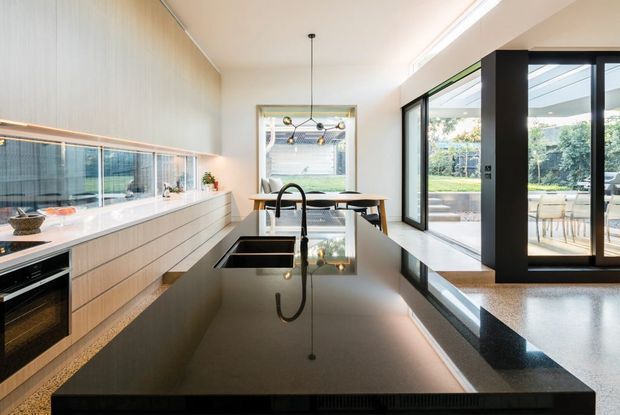 A bright contemporary kitchen with a large black stone island and integrated sink, pale timber cabinetry, and a long horizontal mirrored or glazed backsplash. Full-height black-framed sliding doors open to an outdoor dining terrace, while a timber dining table and pendant light sit at the far end. The space has clean lines, abundant natural light, and a strong indoor-outdoor connection.