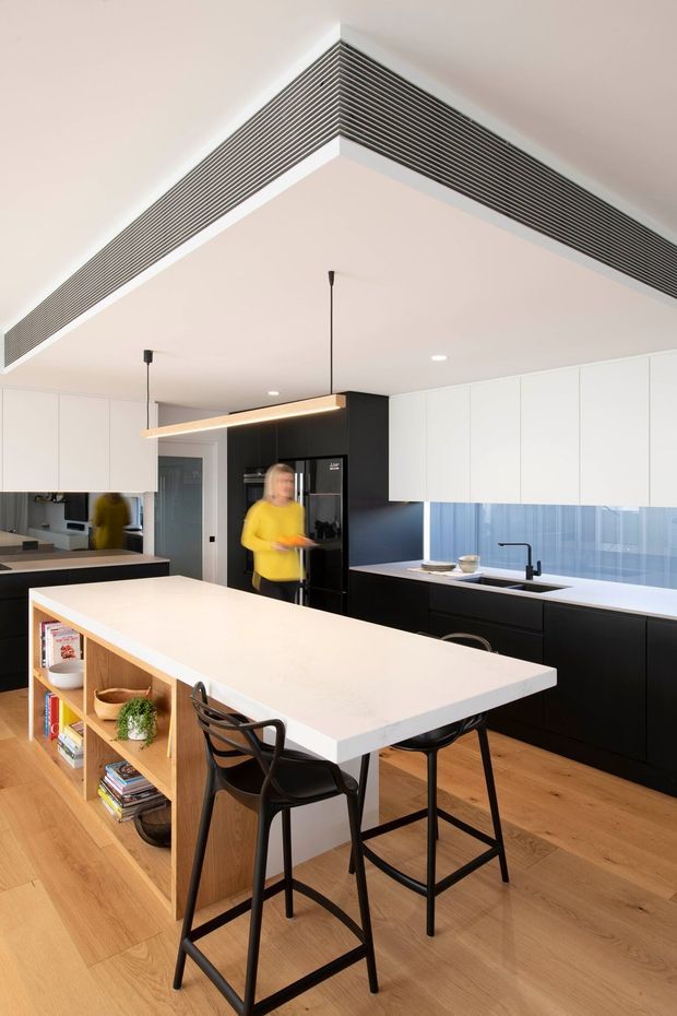 Bright contemporary kitchen interior with a large white island and integrated timber open shelving, black cabinetry, white wall cabinetry, and a dark linear ventilation grille detail wrapping the ceiling bulkhead. The island includes bar seating, while timber flooring, pendant lighting, and a blue-toned tiled or glass splashback complete the space.