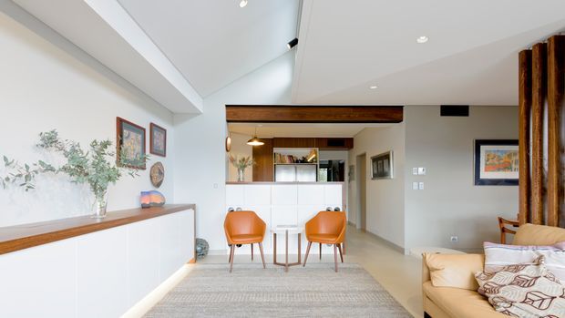 Bright contemporary open-plan living and dining area with a white built-in sideboard, warm timber detailing, exposed timber beam, compact dining table with two orange chairs, and a beige sofa. Recessed ceiling lighting and clean-lined cabinetry create a light, refined interior, with artwork and indoor greenery adding colour and softness.